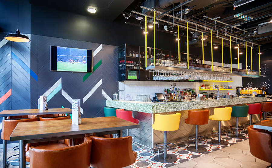 Modern bar featuring wooden tables and tan recliner seats with a video display on the left showing a soccer game and a full bar on the right.