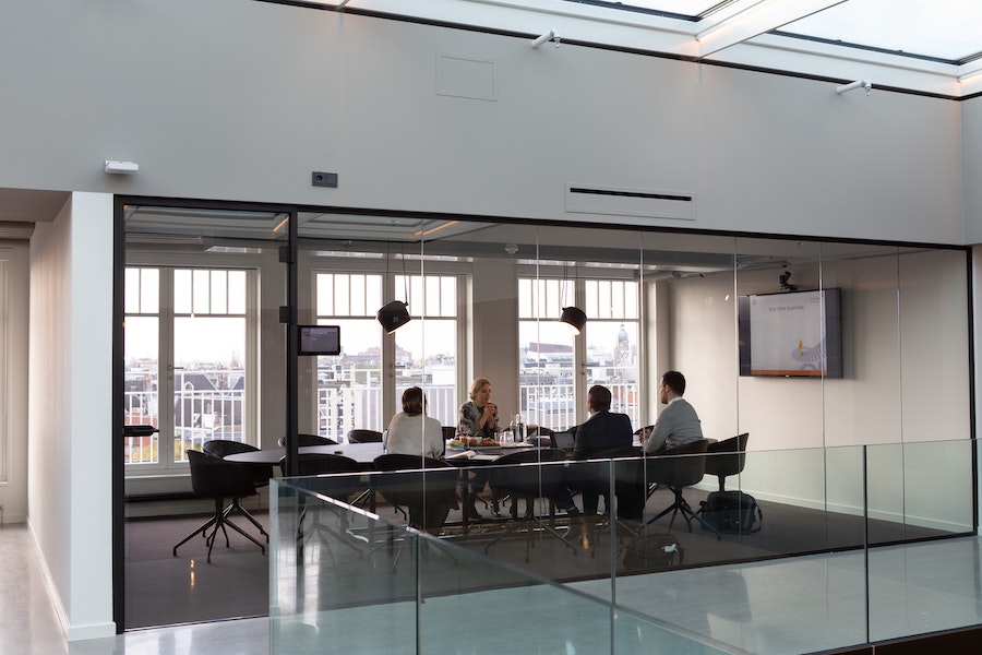 Glass-walled conference room featuring overhead lights, TV display, and four employees around a meeting table.